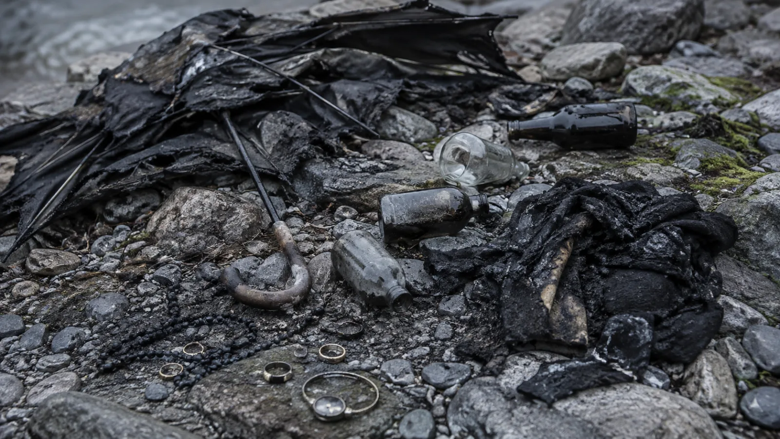 Illustrative still life of scattered burned objects on rocky ground, including charred fabric, an umbrella, glass bottles, and jewelry